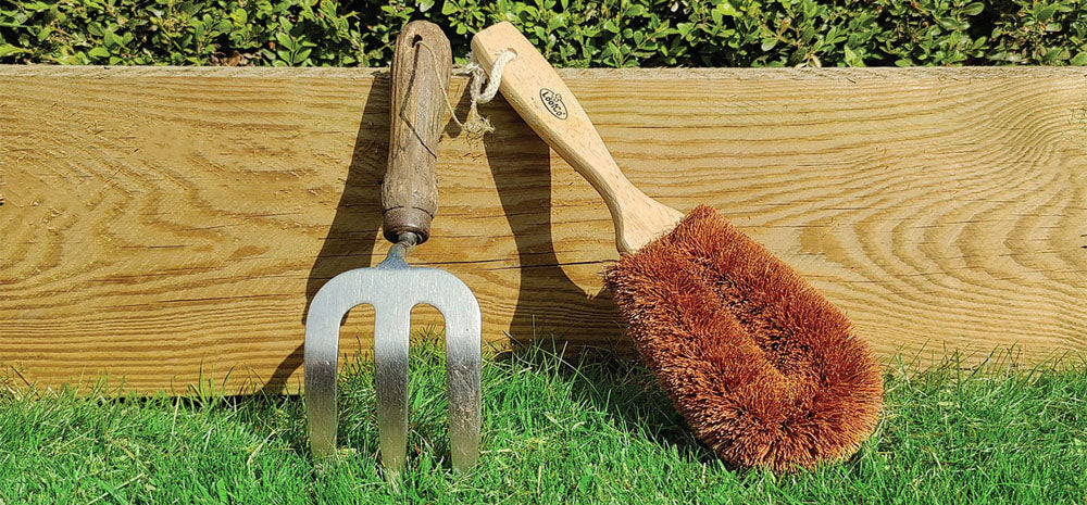 LoofCo Gardeners Brush and small garden fork against a wooden background with grass in foreground