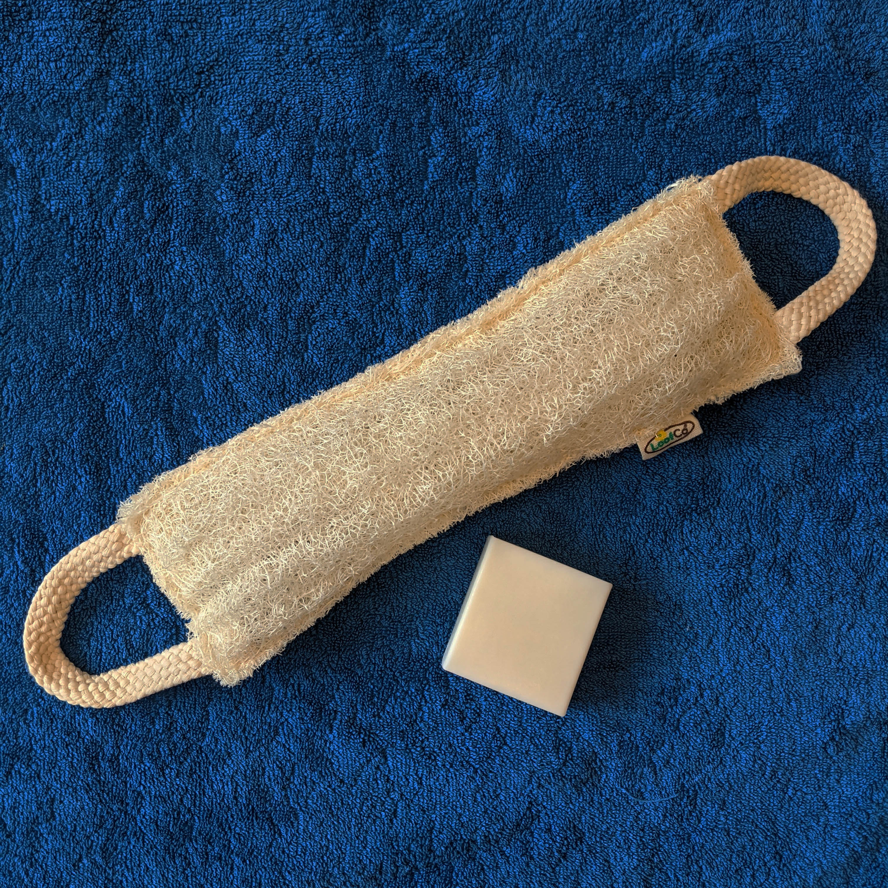 LoofCo back Scrubber made from loofah with a bar of saop on a blue towel
