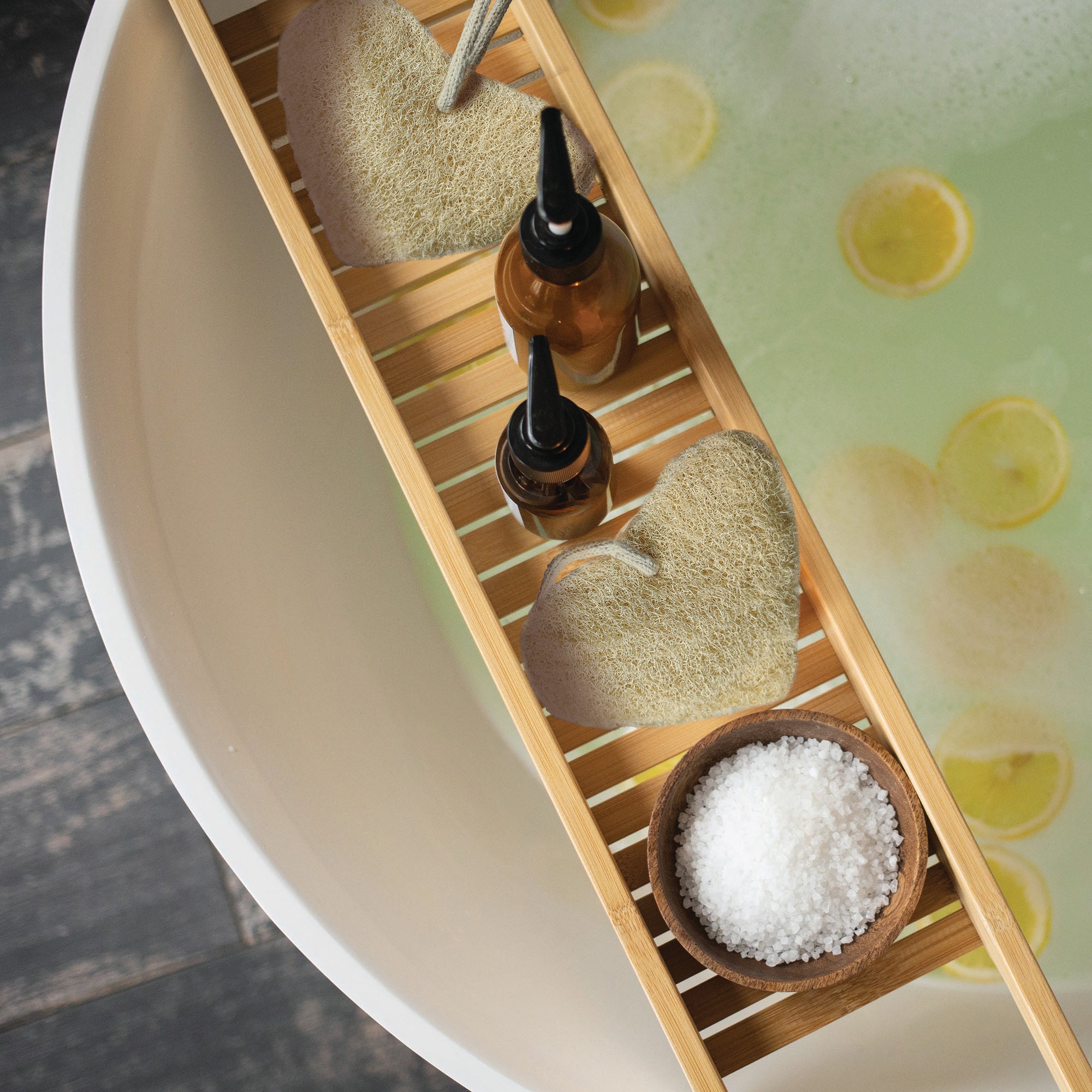 Bath caddy with loofahs, bottles, and a bowl of salt next to a bathtub filled with water and lemons.