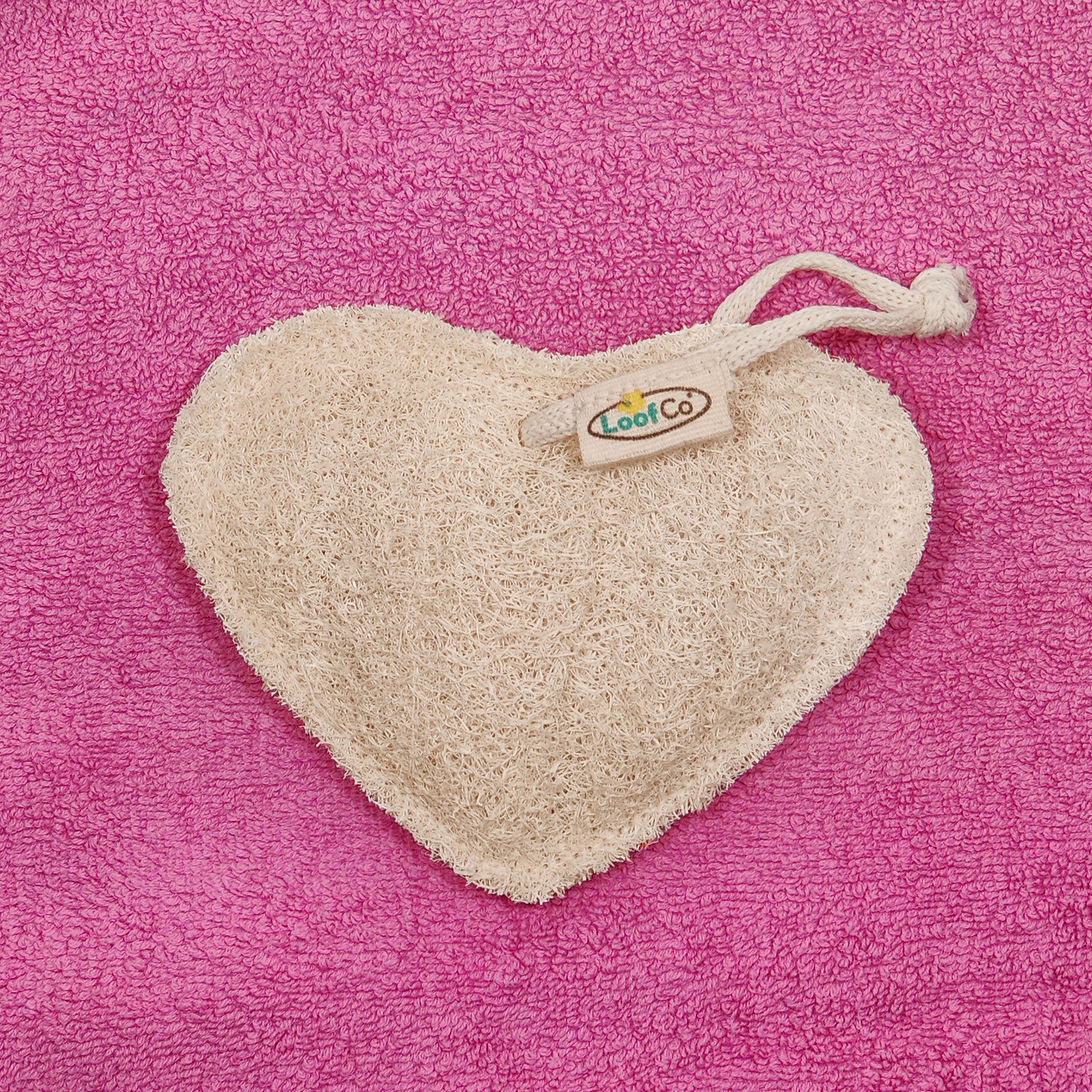 Heart-shaped LoofCo Bath-Time exfoliating pad with a brand tag on a pink textured background