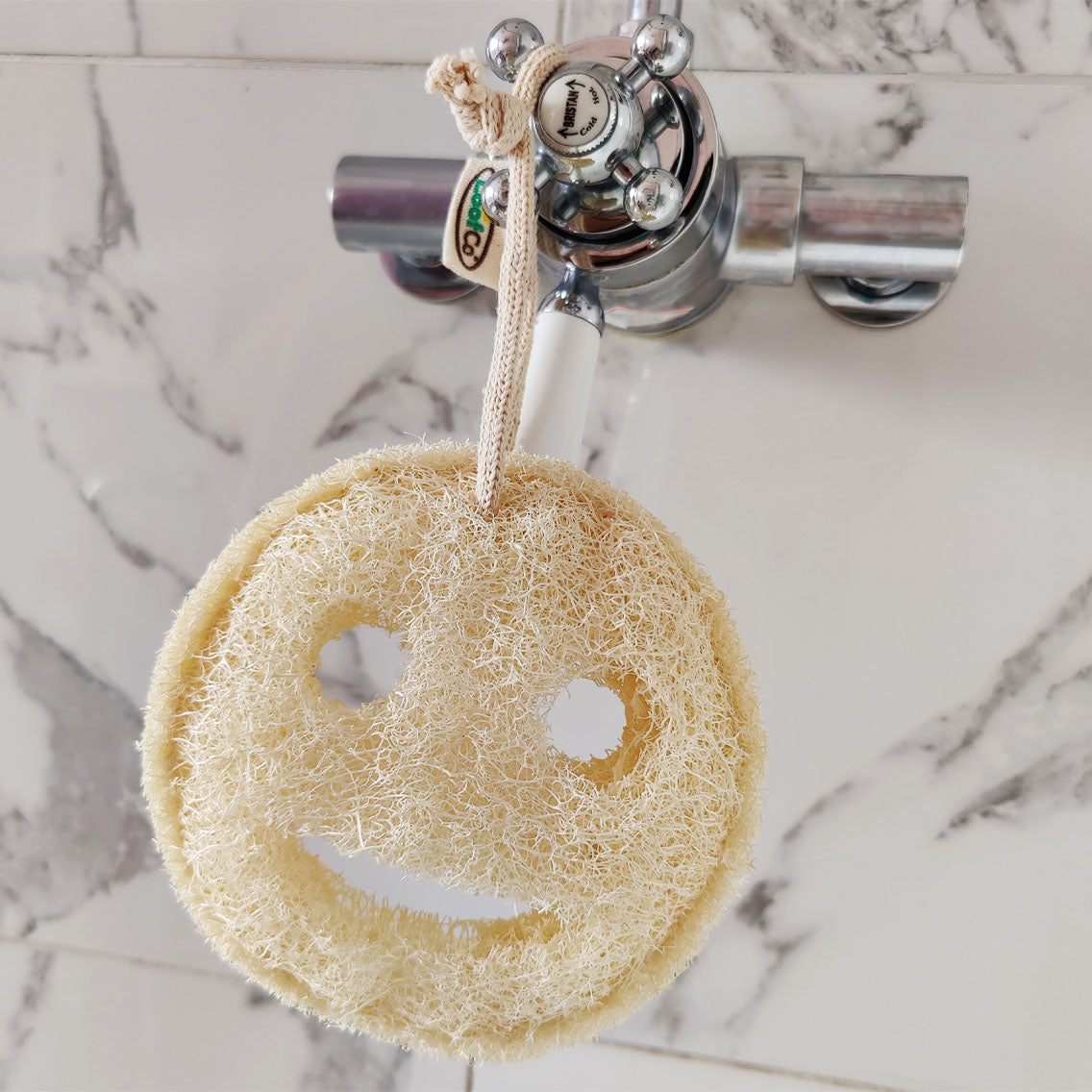 Smiley face loofah hanging on a shower handle against a marble background