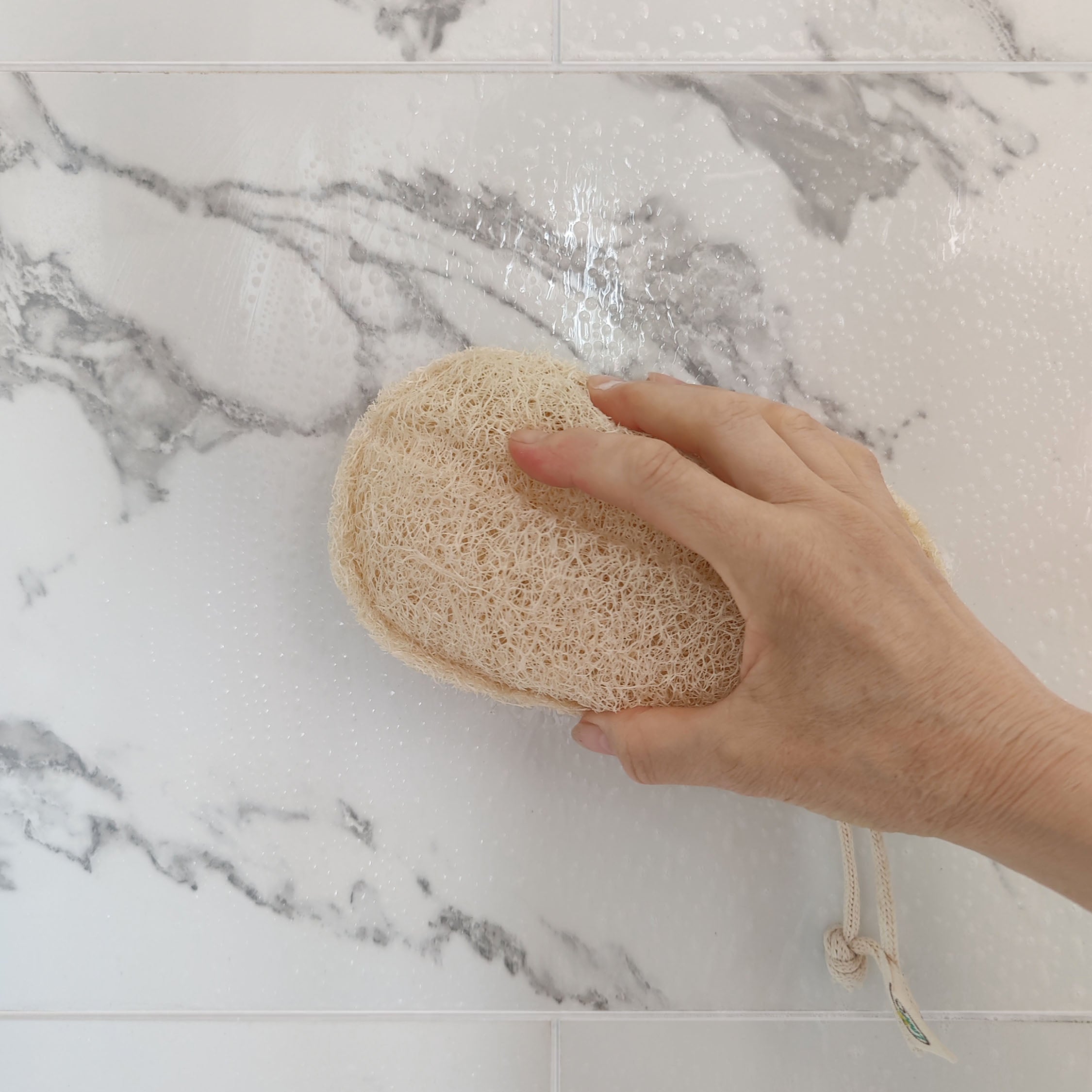 Hand holding a natural LoofCo Bathroom Cleaning pad loofah sponge against a marble bathroom background