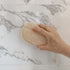 Hand holding a natural LoofCo Bathroom Cleaning pad loofah sponge against a marble bathroom background
