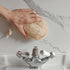 Hand holding a LoofCo bathroom Cleaning Pad loofah sponge above a tap on a marble countertop