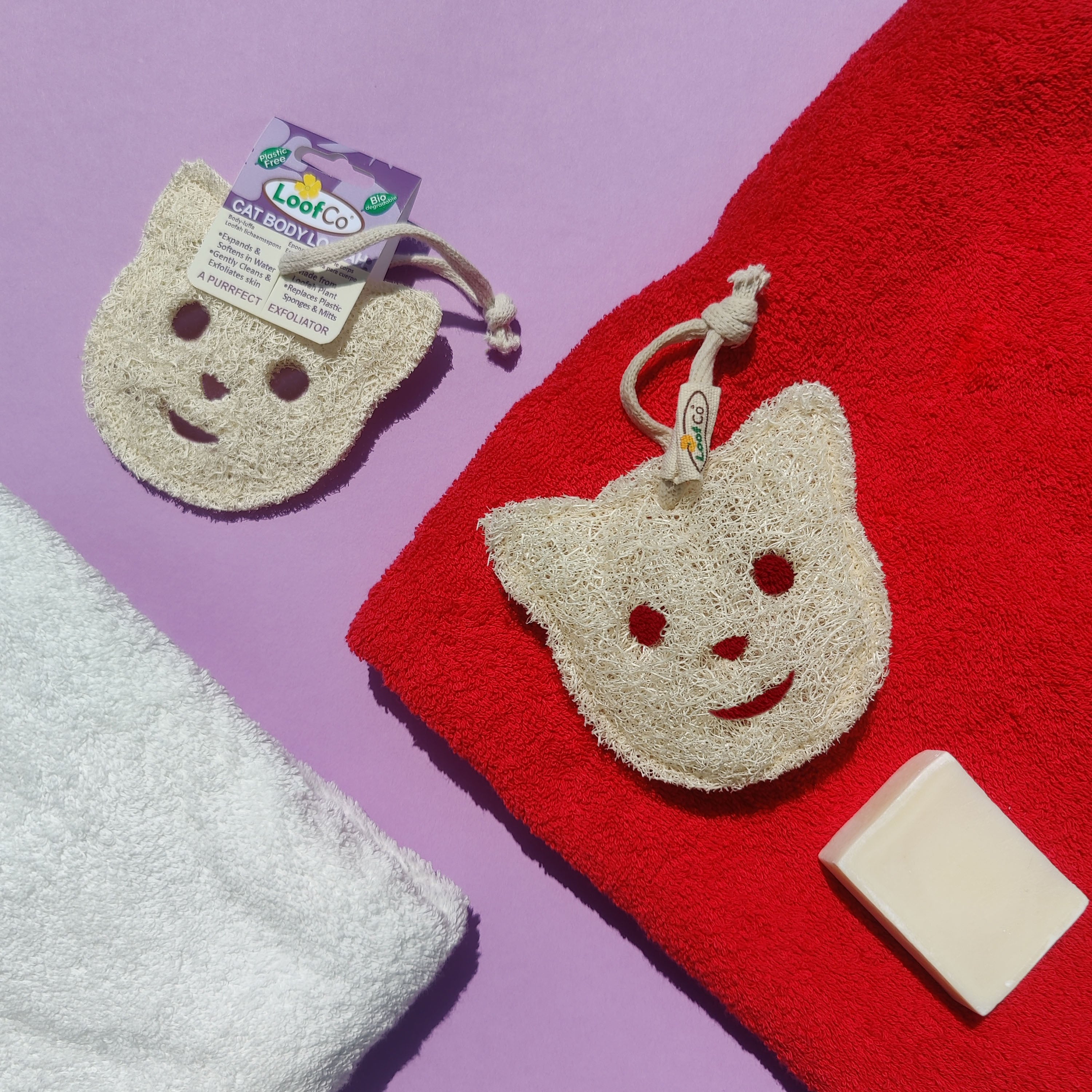 LoofCo Cat-shaped bath pads with a smiley face on a red towel with soap, on a purple background.