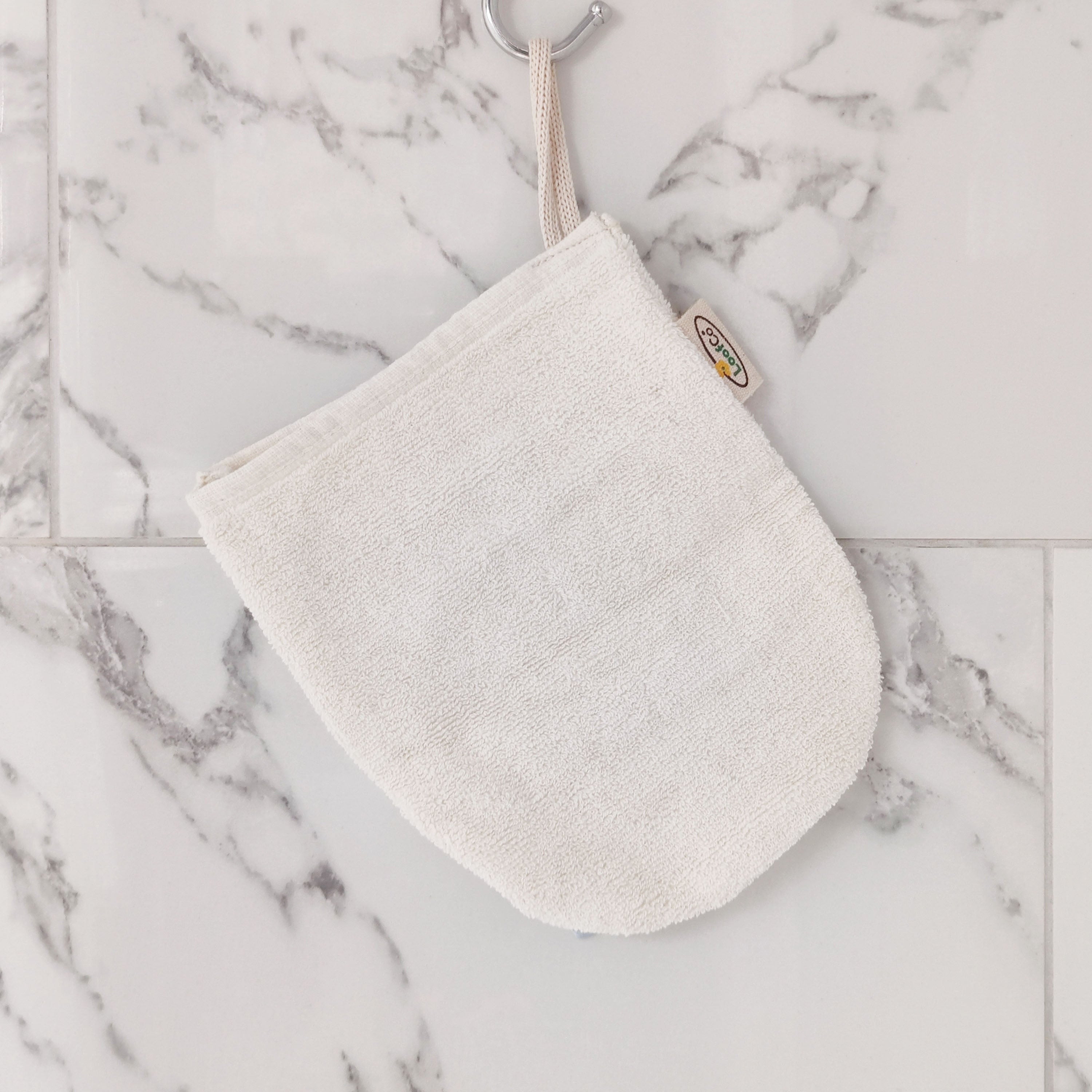 LoofCo Cleansing Mitt hanging from hook against marble tiled shower cubicle