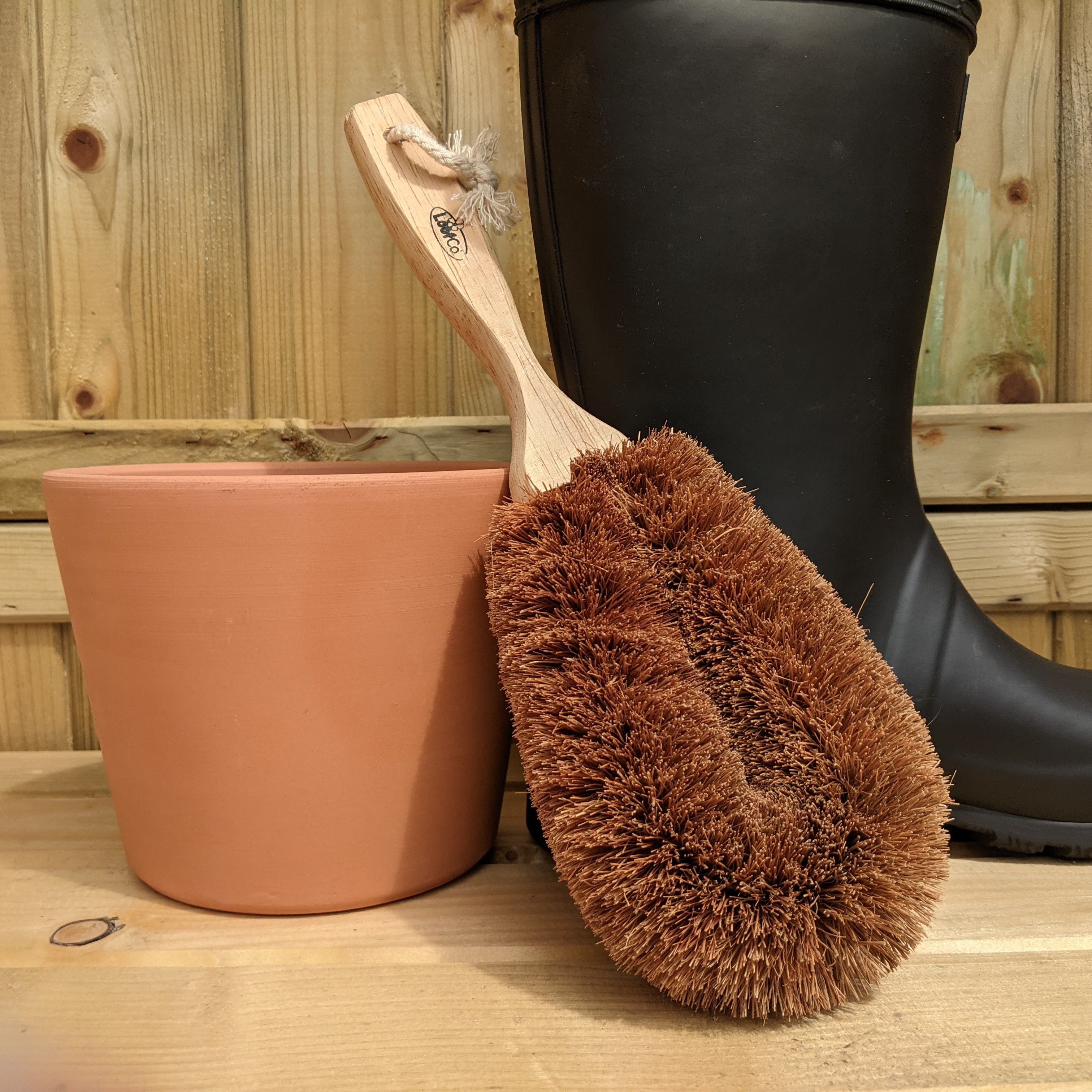 LoofCo Coir Gardener's Brush with wooden handle next to a black rubber boot and terracotta pot on a wooden surface.