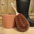 LoofCo Coir Gardener's Brush with wooden handle next to a black rubber boot and terracotta pot on a wooden surface.