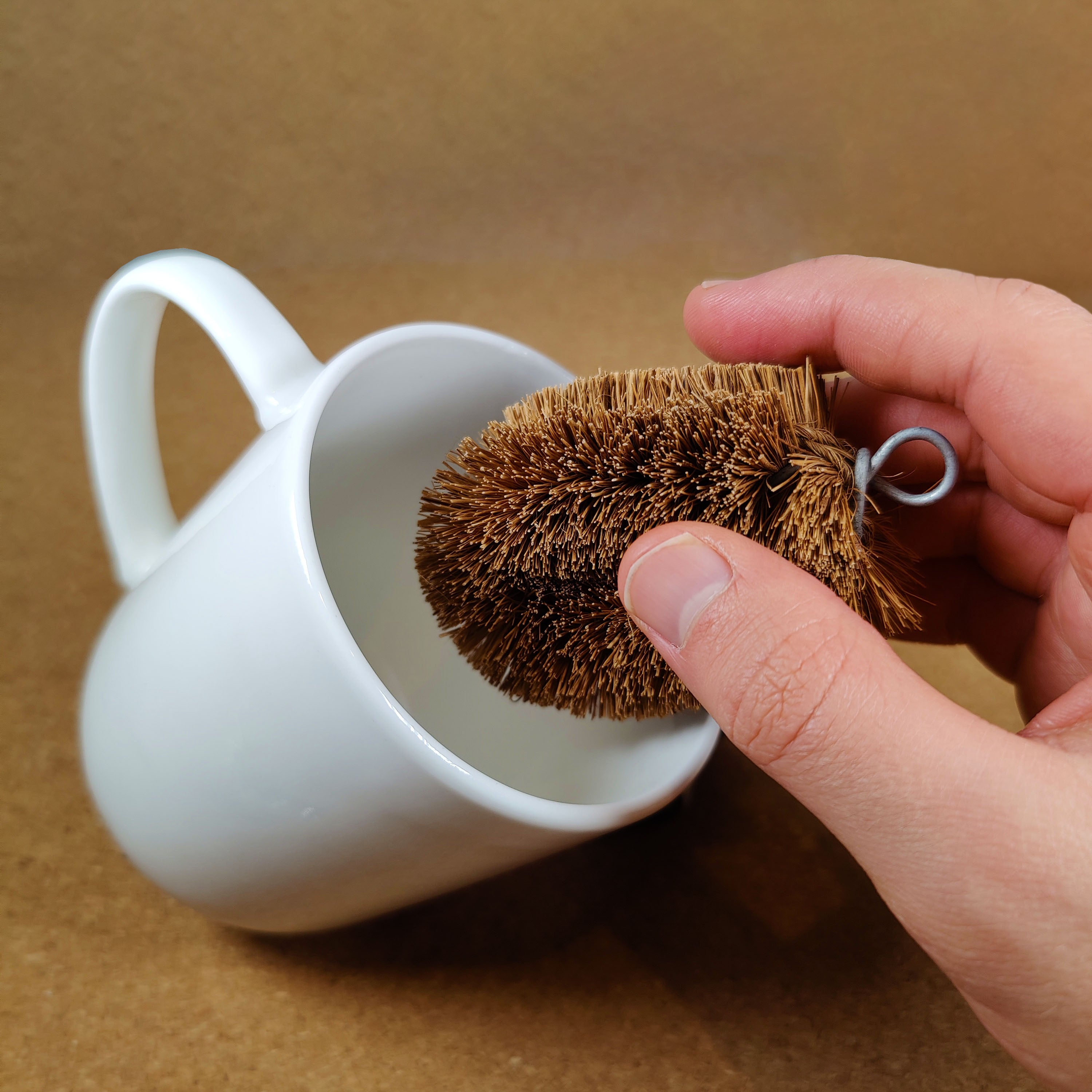 Hand holding a brown LoofCo Mini Scrub Brush near a white mug on a brown background