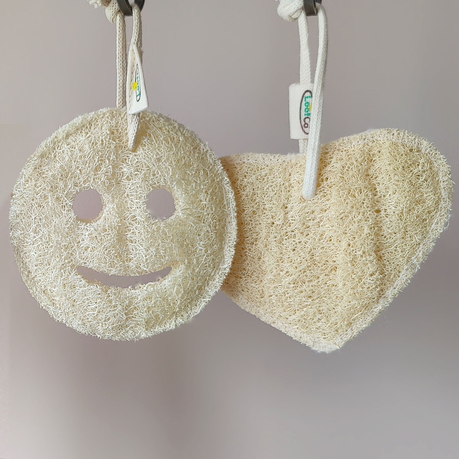 Two natural LoofCo loofahs, one round with a smiley face and one heart-shaped, hanging against a neutral background.