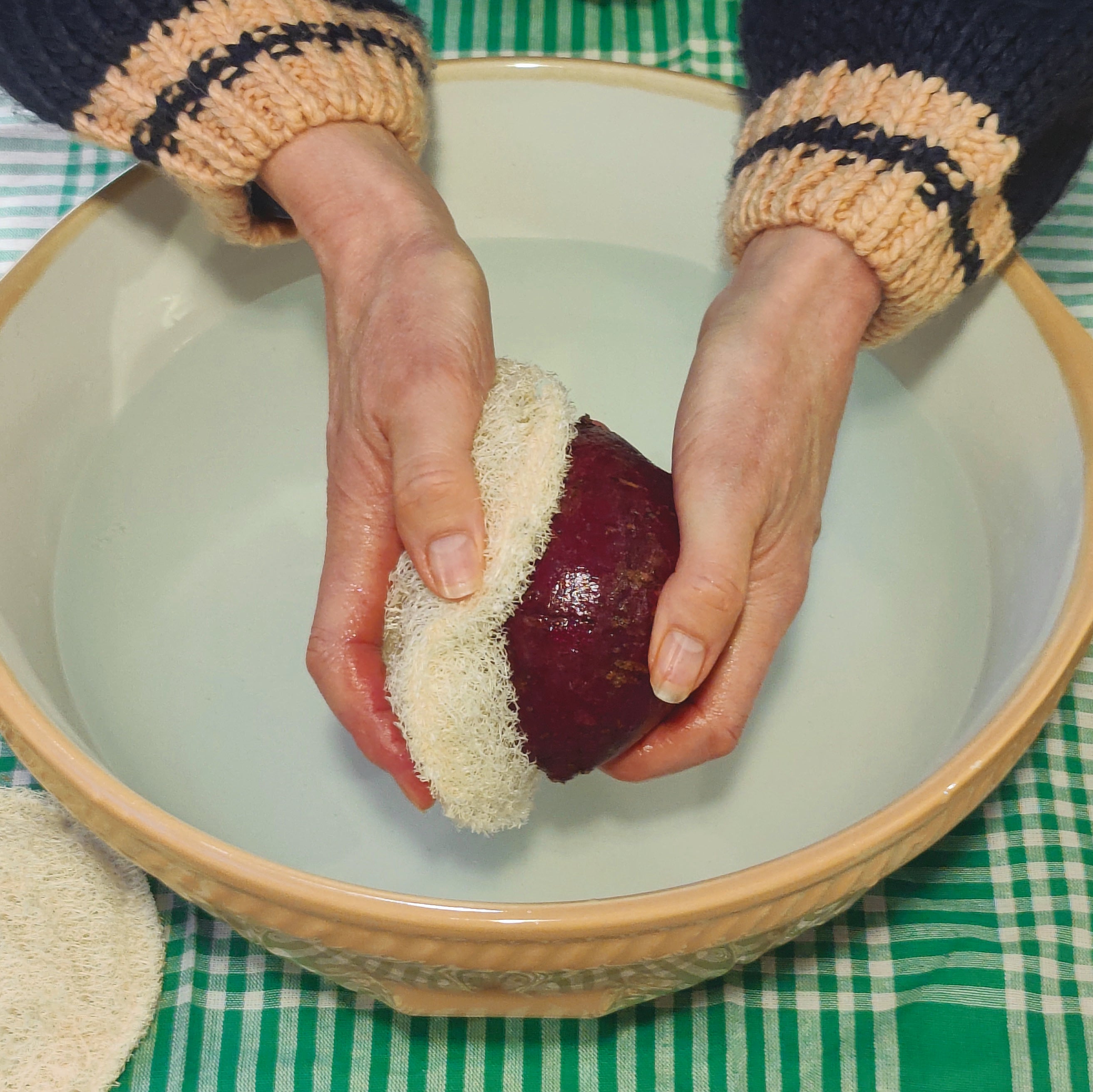 Person scrubbing a sweet potato with a Loofah root vegetable scrubber over a  bowl on a green checked tablecloth.