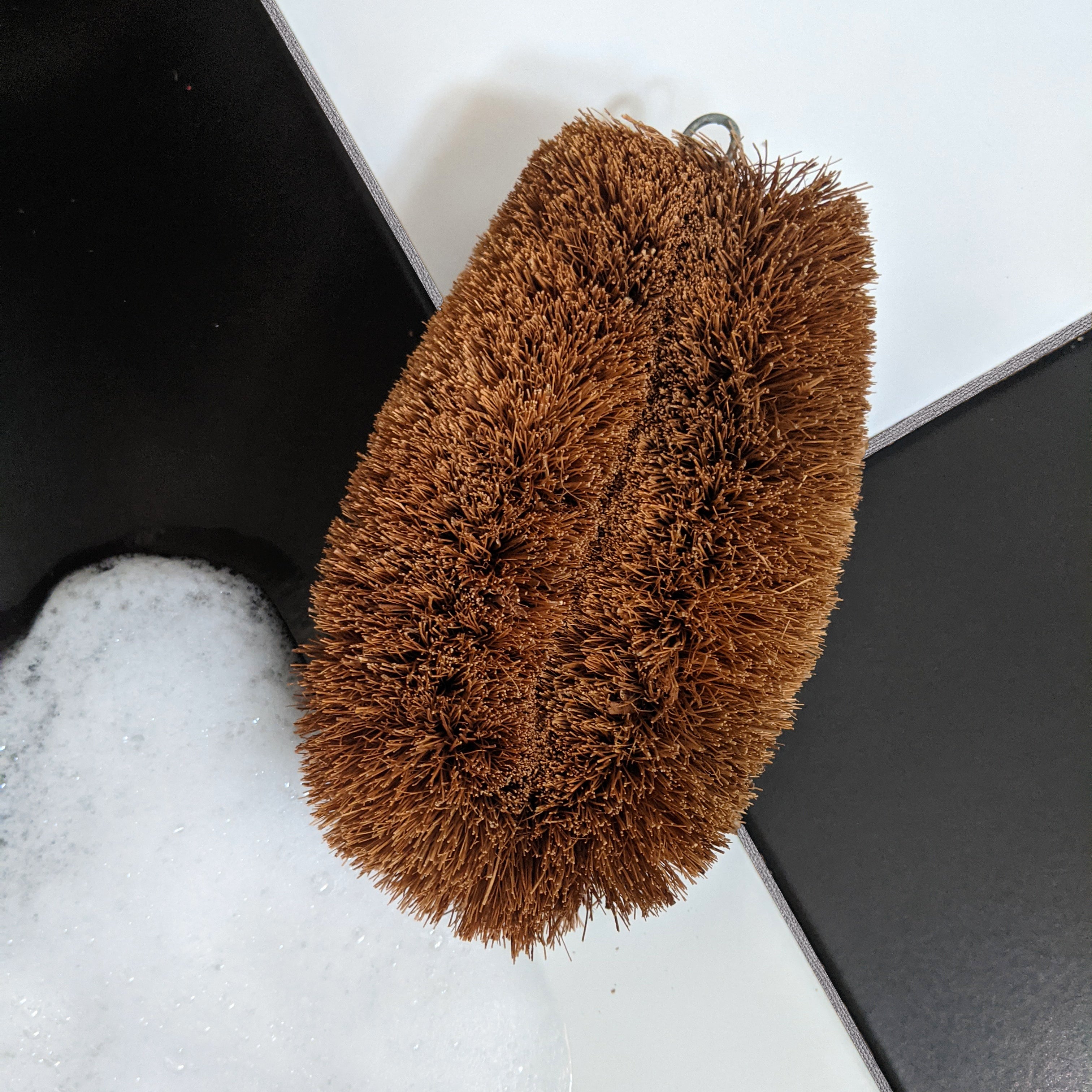 LoofCo Scrubbing Brush on a white surface with a black object and soapy water.