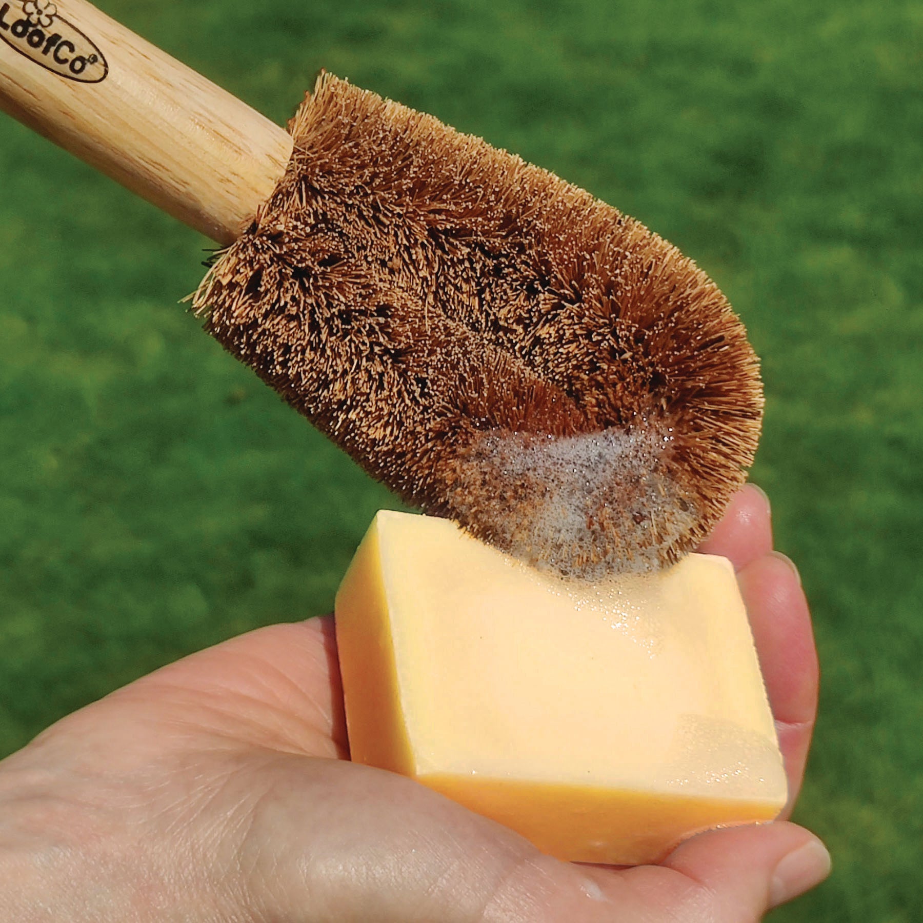 Person holding a LoofCo washing-Up Brush with coir bristles against a lemongrass Washing-Up Soap Bar on a green background