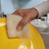 Hand holding a sponge over a yellow plate with soap suds, likely in a kitchen setting.